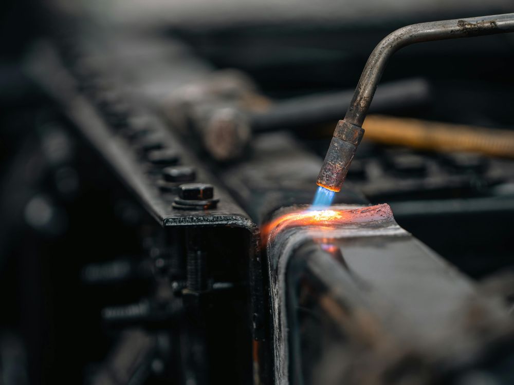 Maçarico aquecendo peça metálica em processo industrial de fabricação do metal, destacando etapa de soldagem e tratamento térmico.